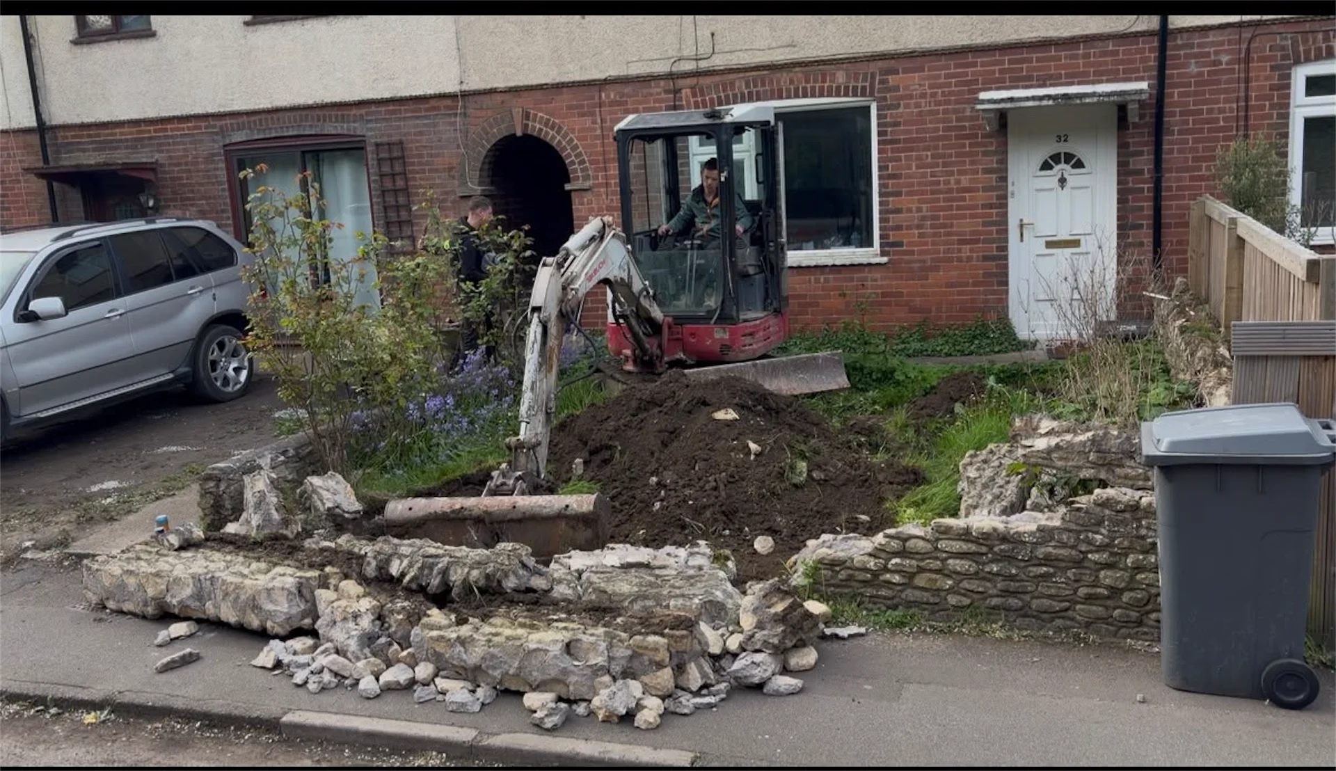 Mini digger excavating old garden wall and ground ready for new driveway installation, Trowbridge