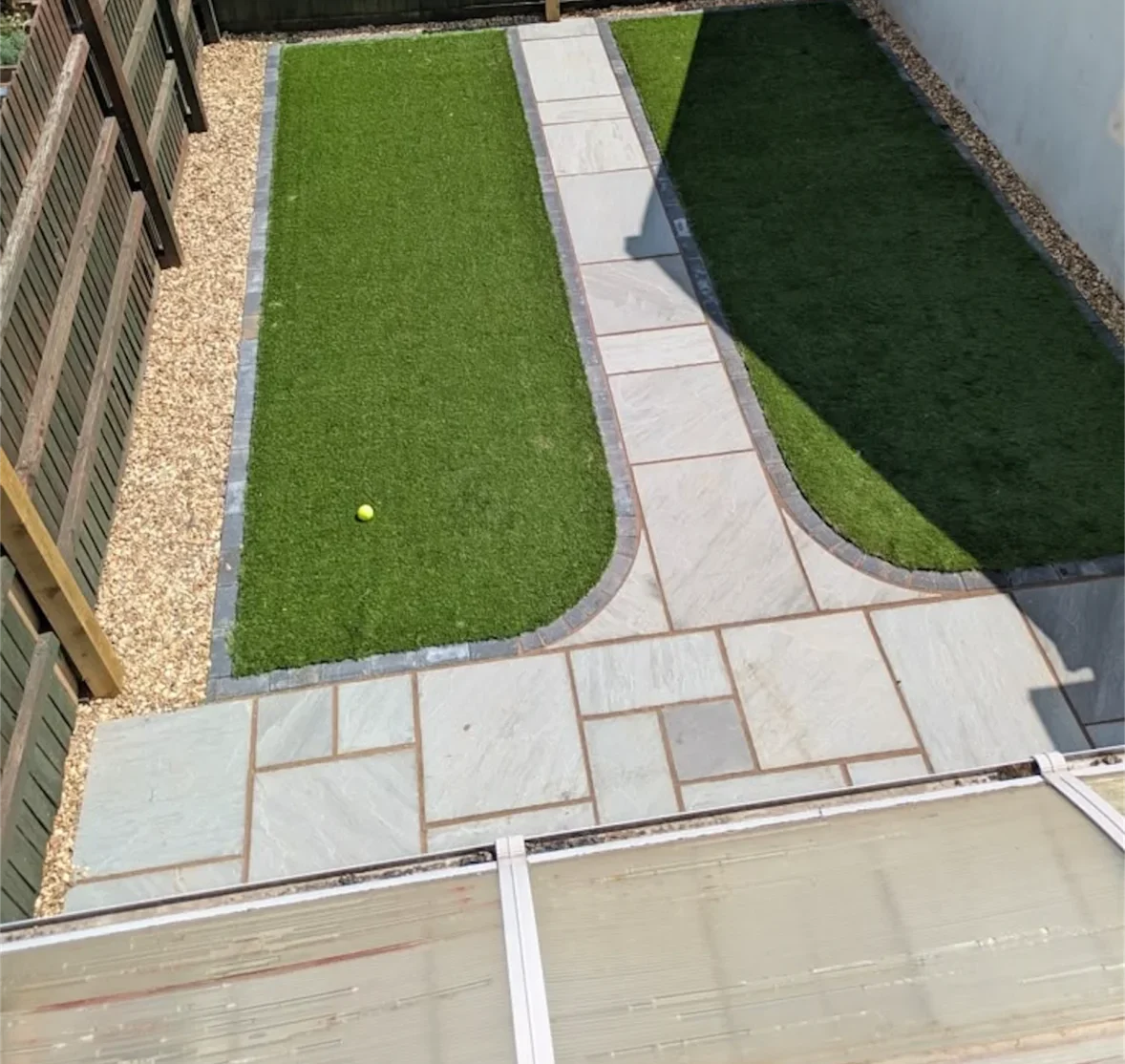 Rear garden with a curved sandstone paved path running between two symmetrical artificial grass lawns, bordered by gravel and charcoal edging
