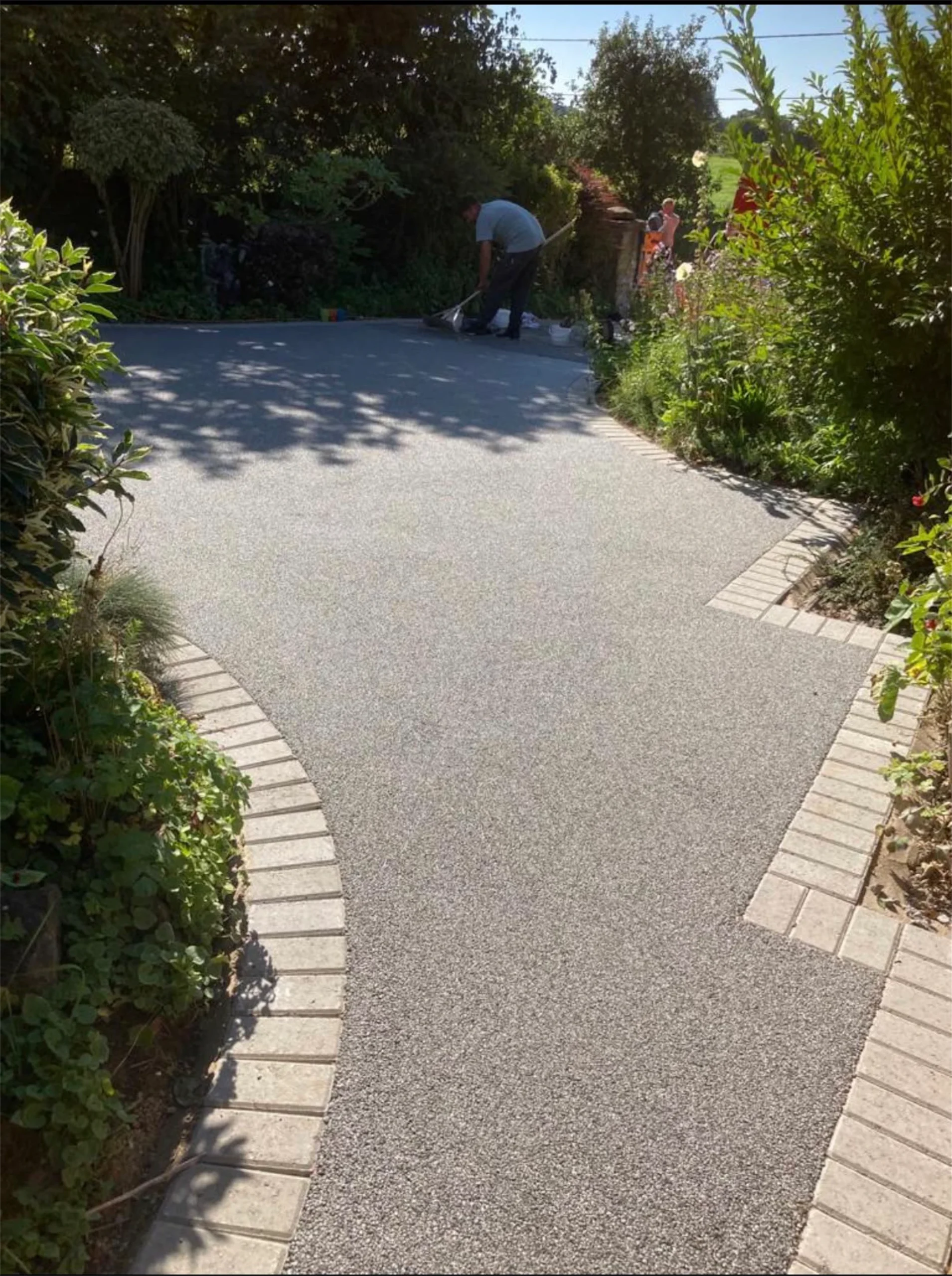 Curved grey resin bound driveway with brick edging, worker finishing the surface amid garden greenery