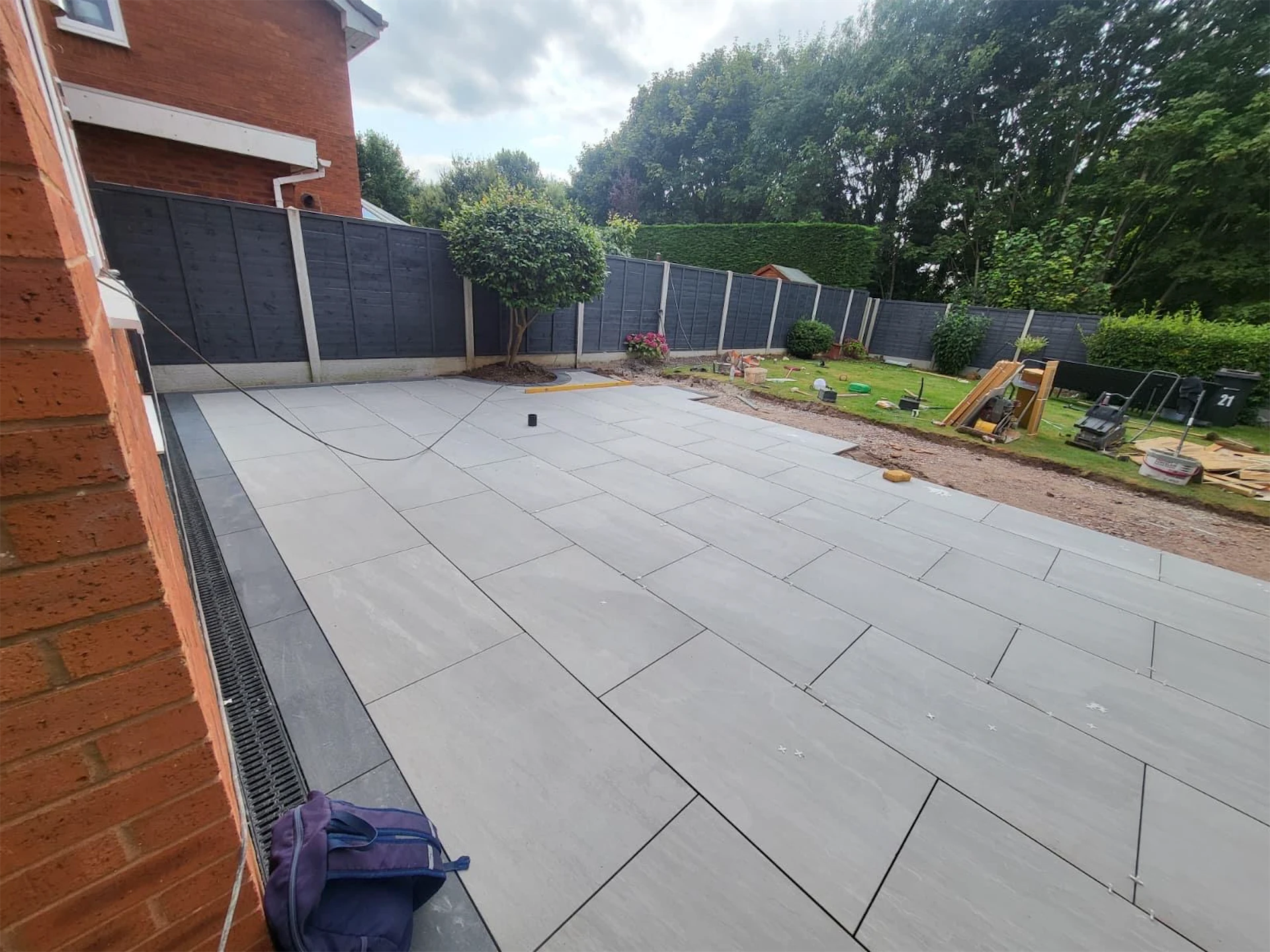 Grey porcelain patio slabs being laid alongside a house, with a linear drainage channel and unfinished garden area